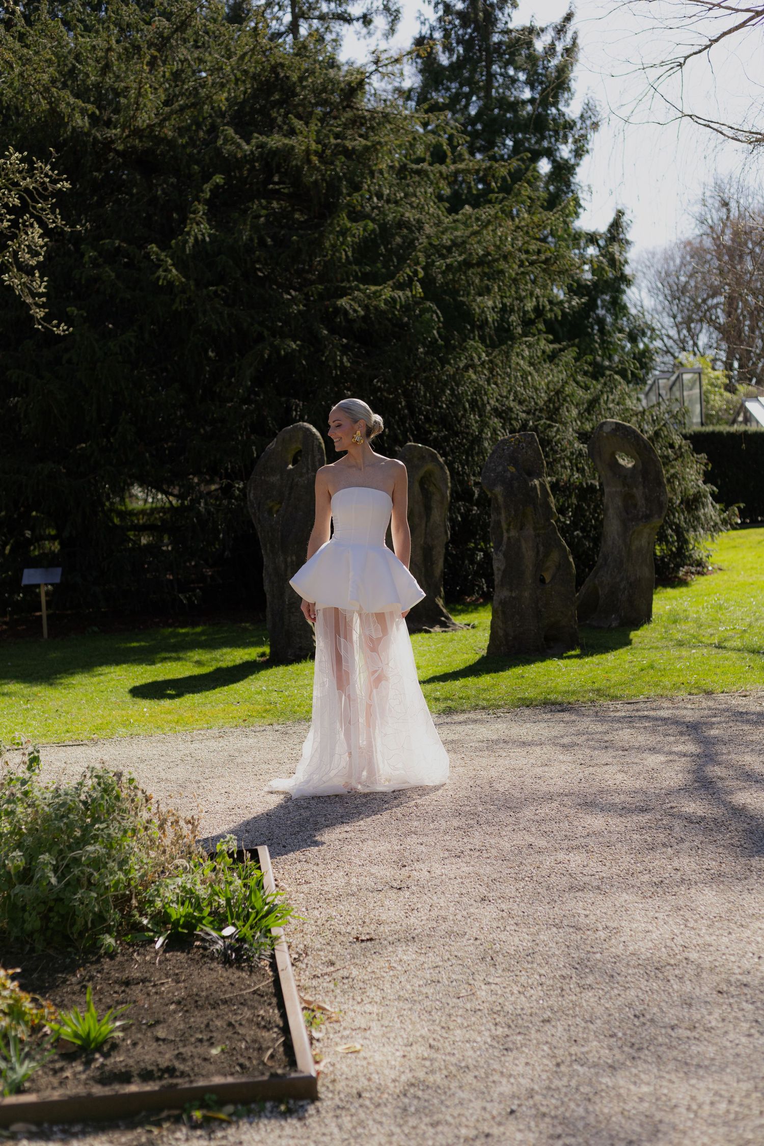 Bride in flowing white dress walking along a garden path surrounded by trees.