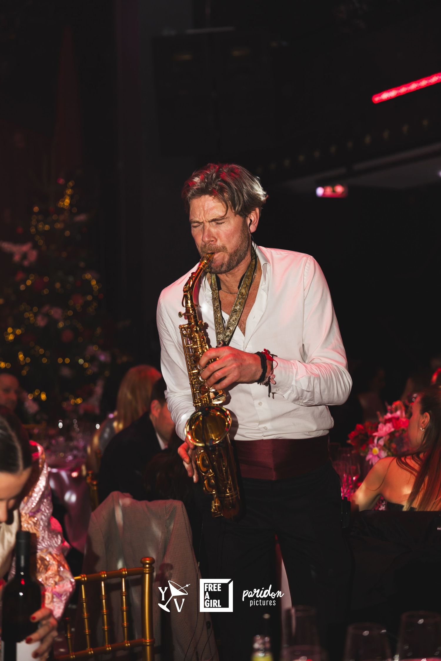 A saxophonist in a white shirt performs passionately on stage during a nighttime event with festive decor.