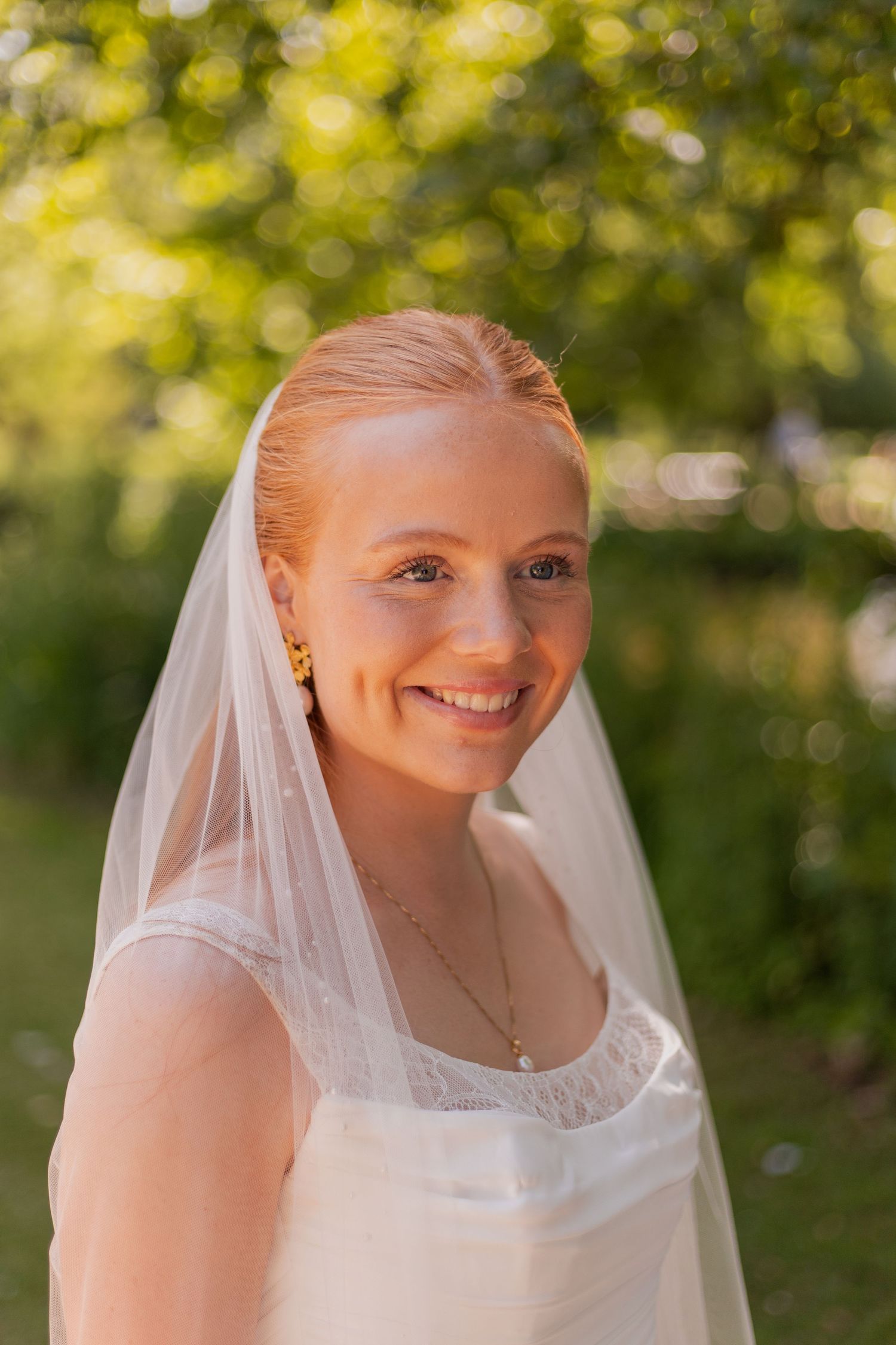 A bride in a white dress and veil smiles radiantly outdoors with green foliage in the background.