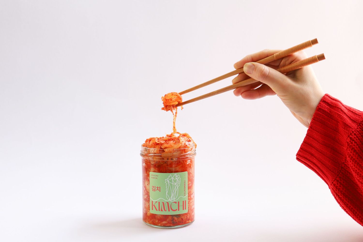 Hands holding chopsticks over an open jar of kimchi.