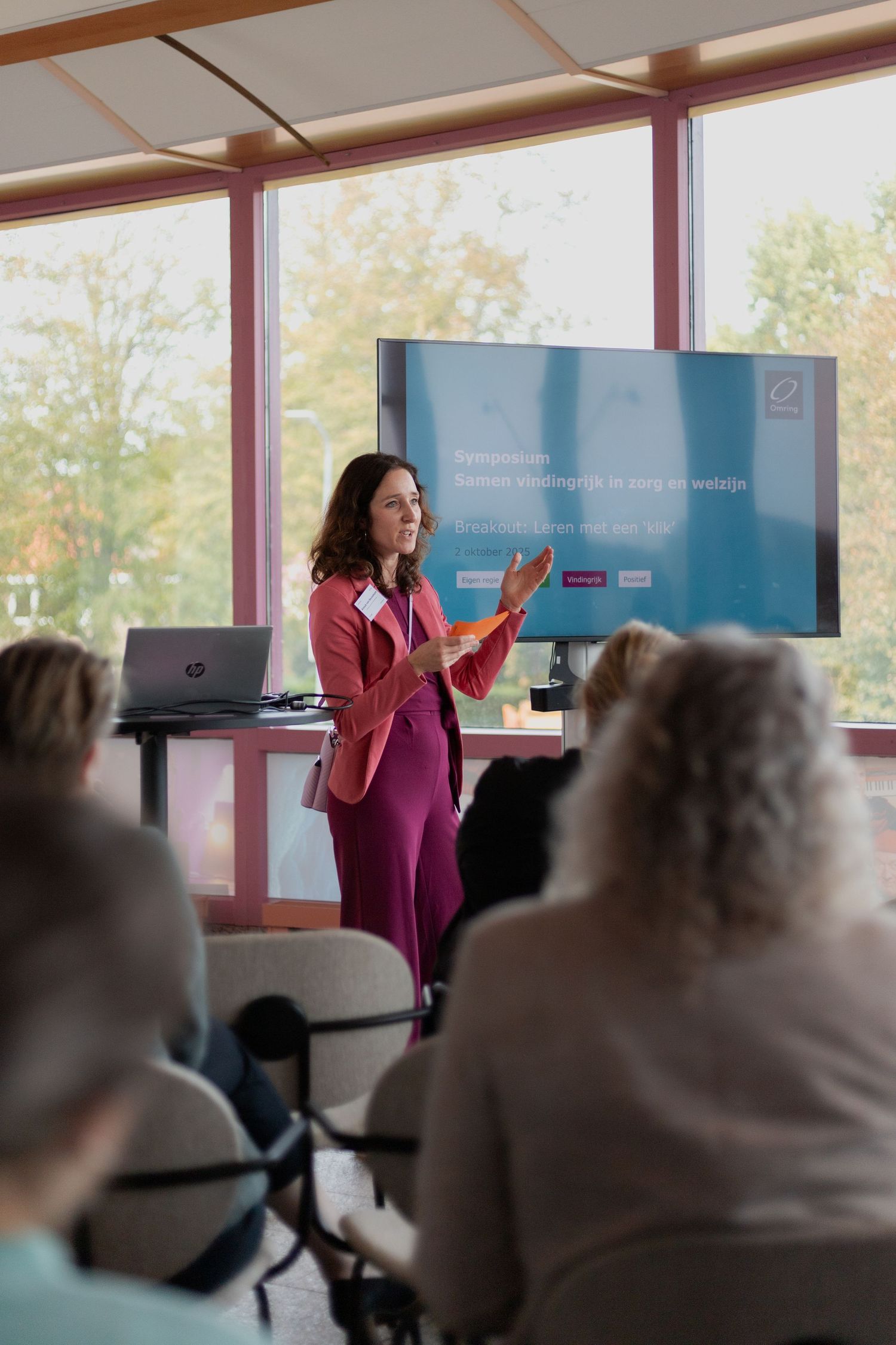 A presenter in a magenta top gives an animated presentation to an audience in a bright conference room with large windows.