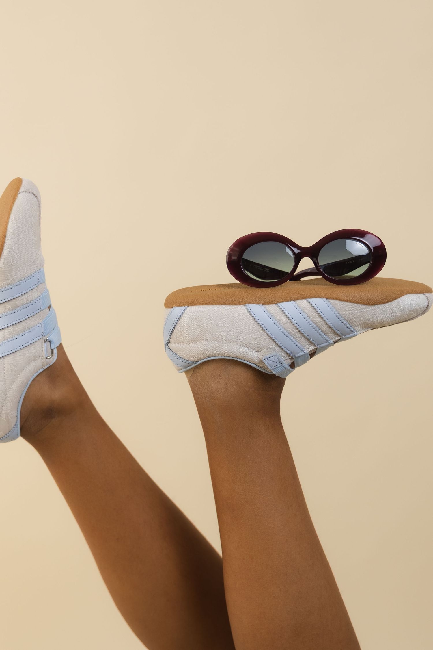 Legs raised wearing light blue Adidas sneakers with a burgundy oval sunglasses resting on the sole against beige background.