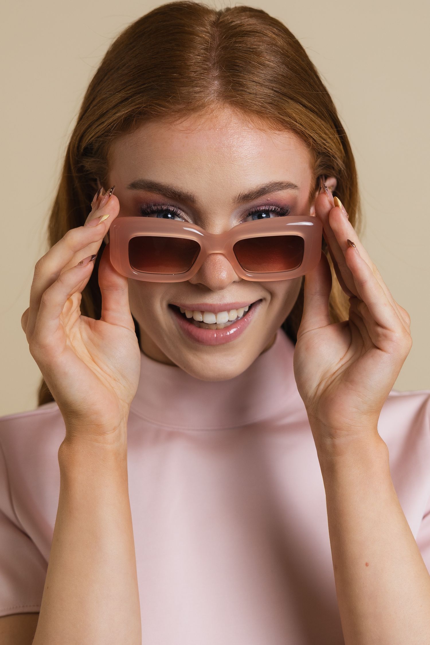 Smiling redhead woman wearing pink rectangular sunglasses and a pink top against a beige background.