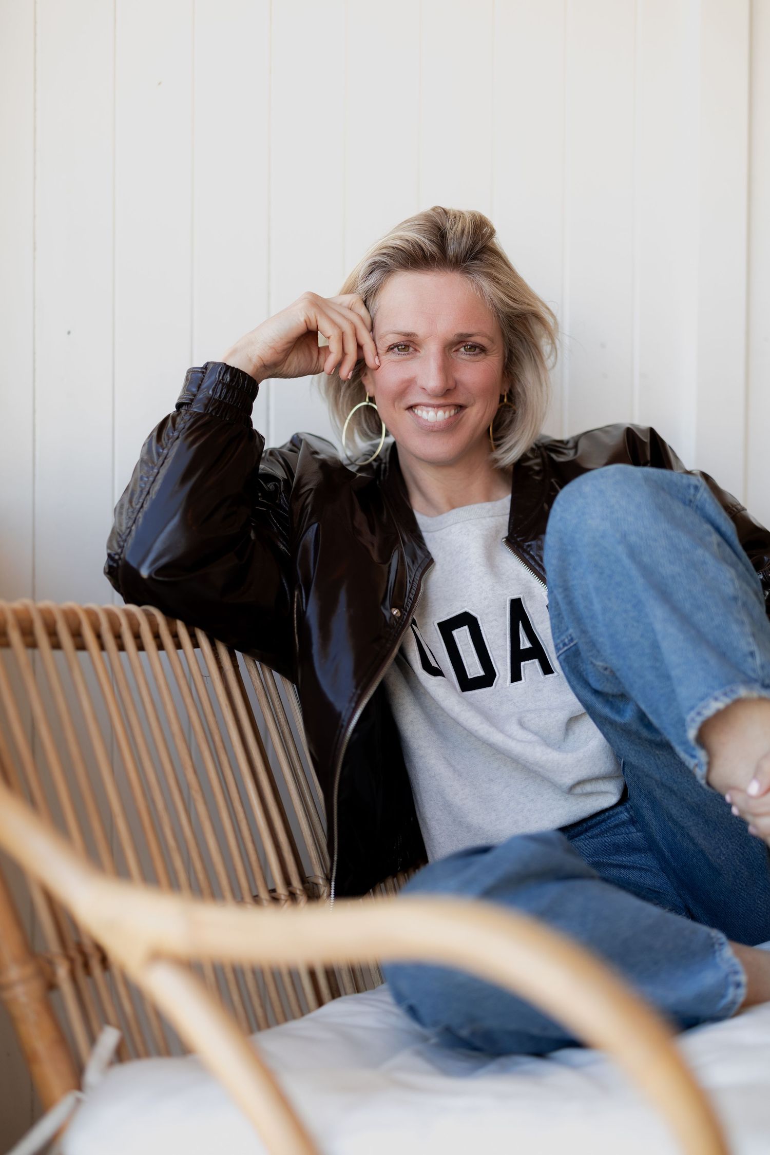 Smiling blonde woman in leather jacket and jeans sitting relaxed on rattan chair against white wall.