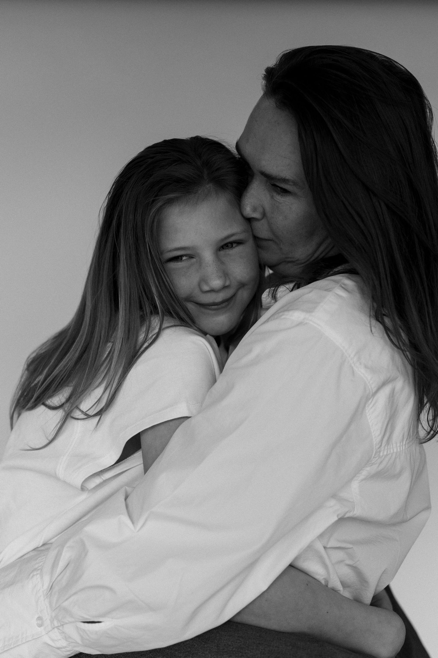 Black and white portrait of a mother kissing her smiling daughter, both wearing white shirts.
