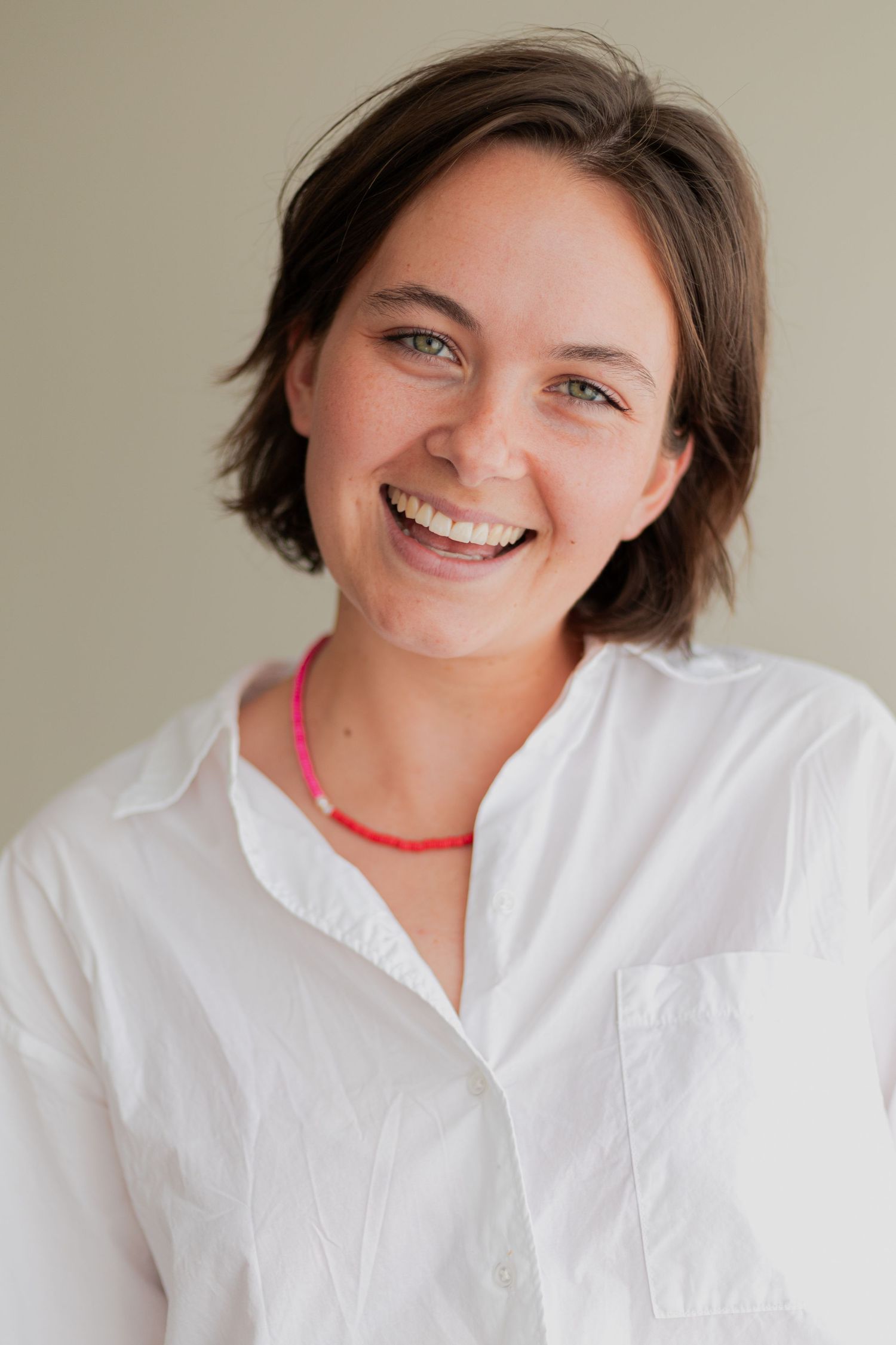 Studio portrait of person in white button-down shirt with red necklace against neutral background.