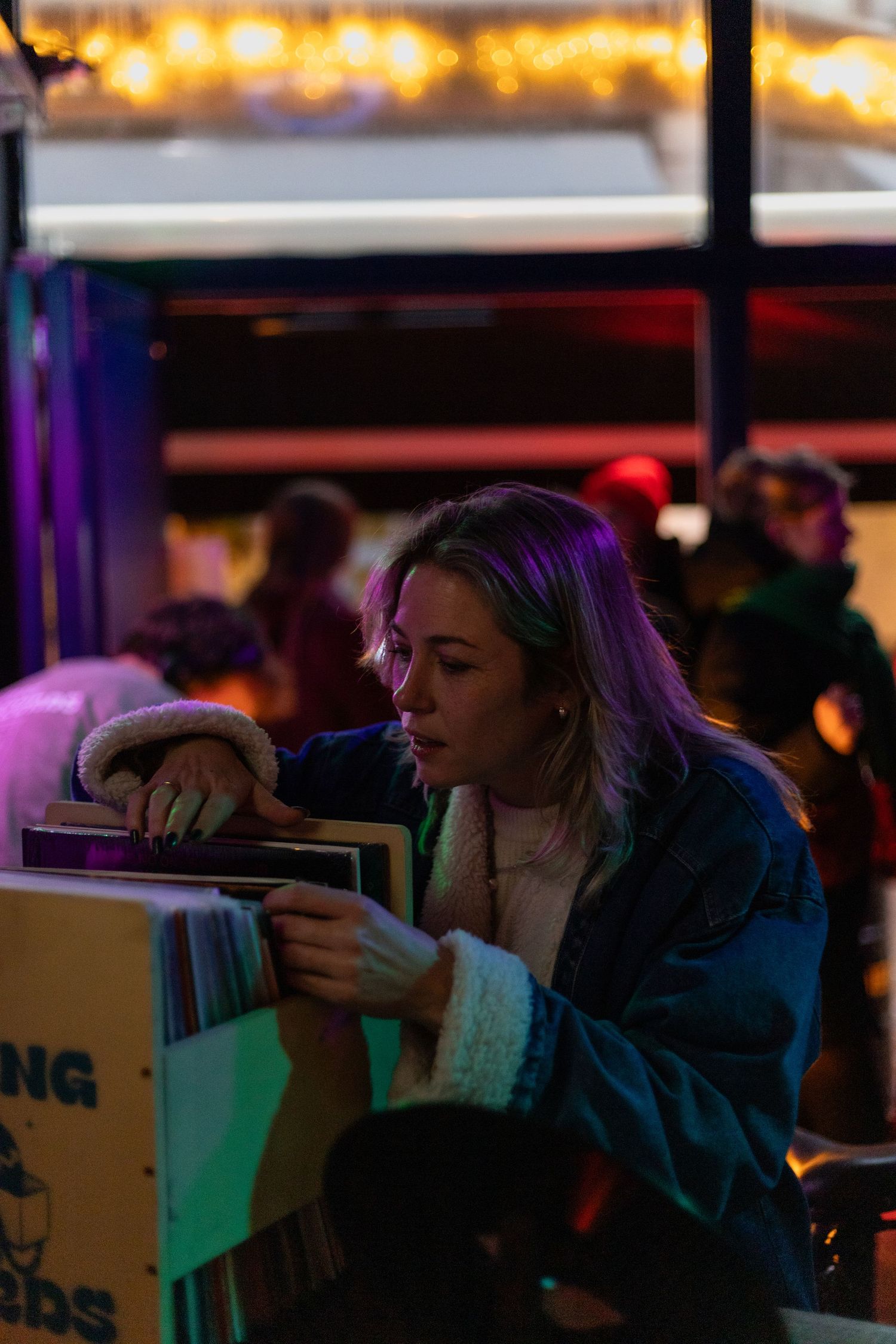 People browse through vinyl records in a dimly lit room with string lights and colorful neon ambiance.