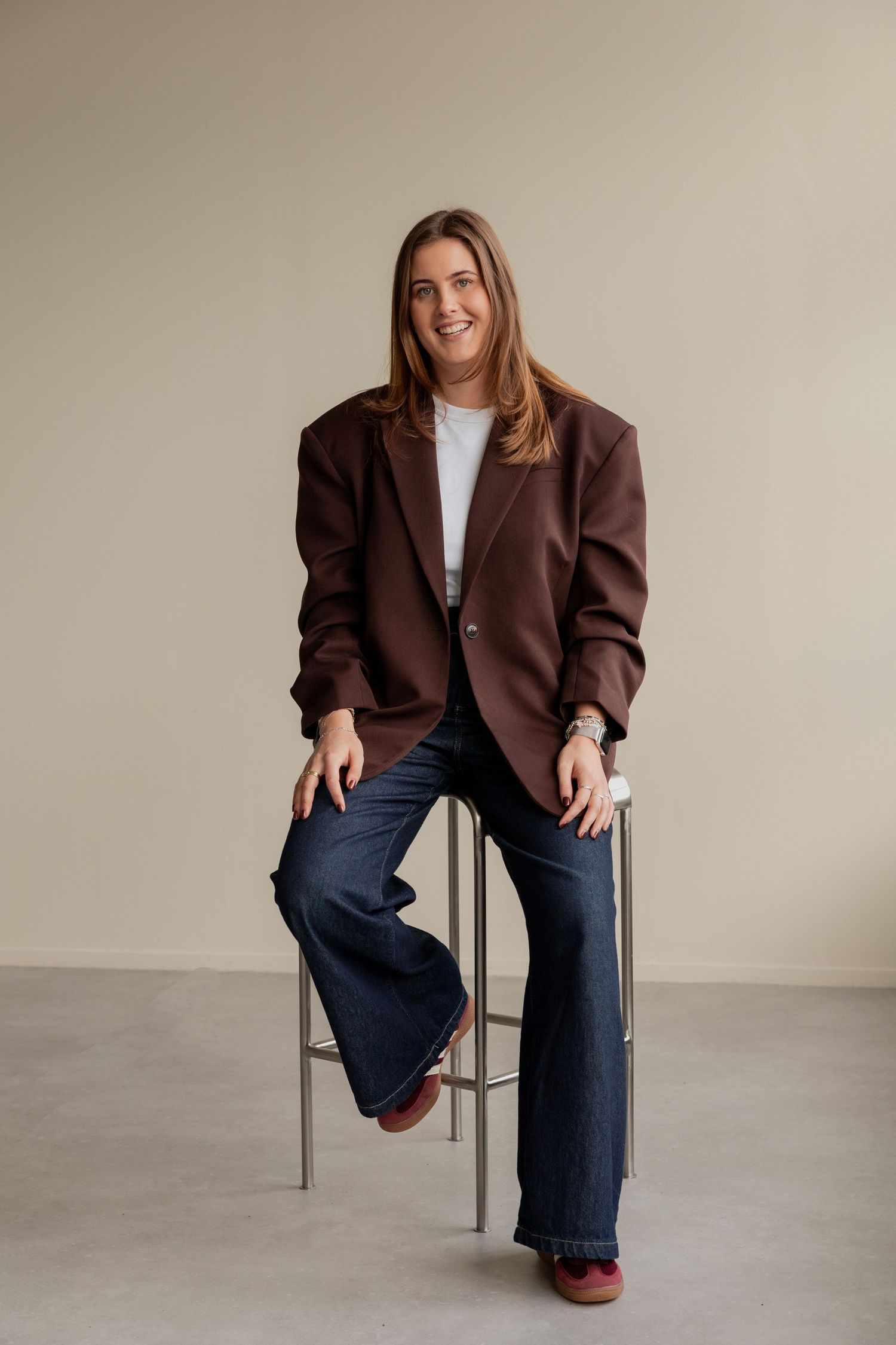Person in brown blazer and jeans sits casually on chair in minimalist setting against neutral wall.