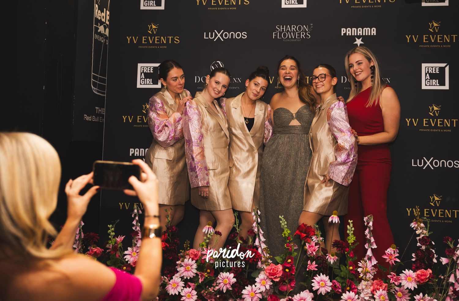 A group of people pose together at a formal event with a branded backdrop displaying luxury logos and text.