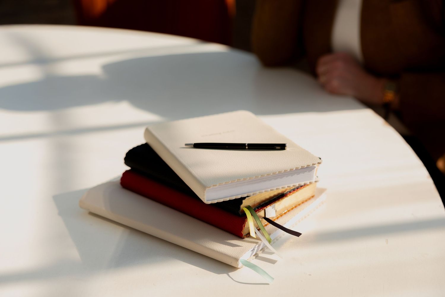Stack of colorful notebooks with a black pen on a white table in warm sunlight.