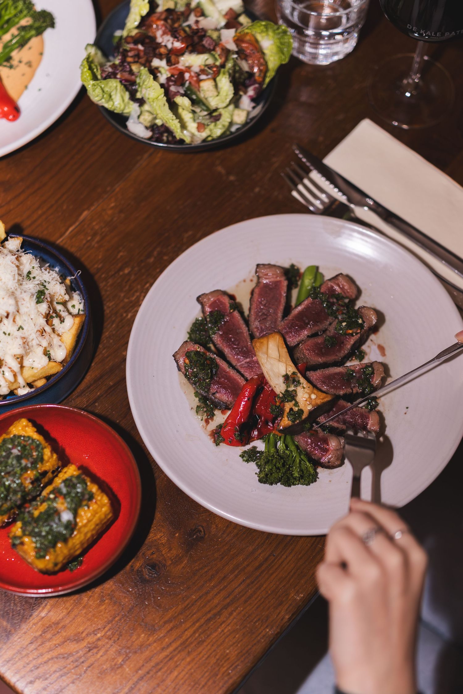 Hands reaching for sliced steak and sides served on white plates at a wooden dining table.