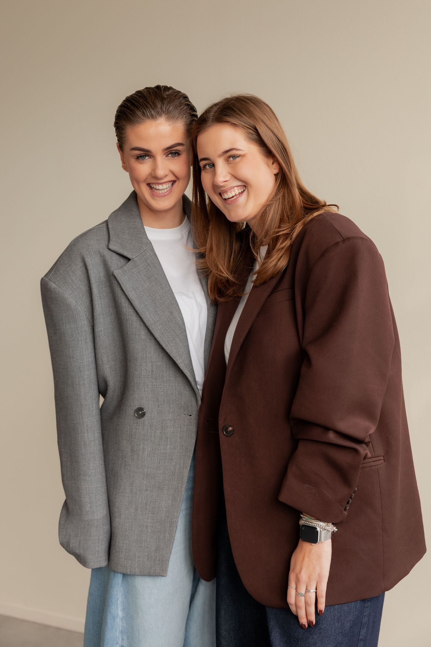 Two people pose together wearing contrasting brown and gray blazers with casual outfits in a studio setting.