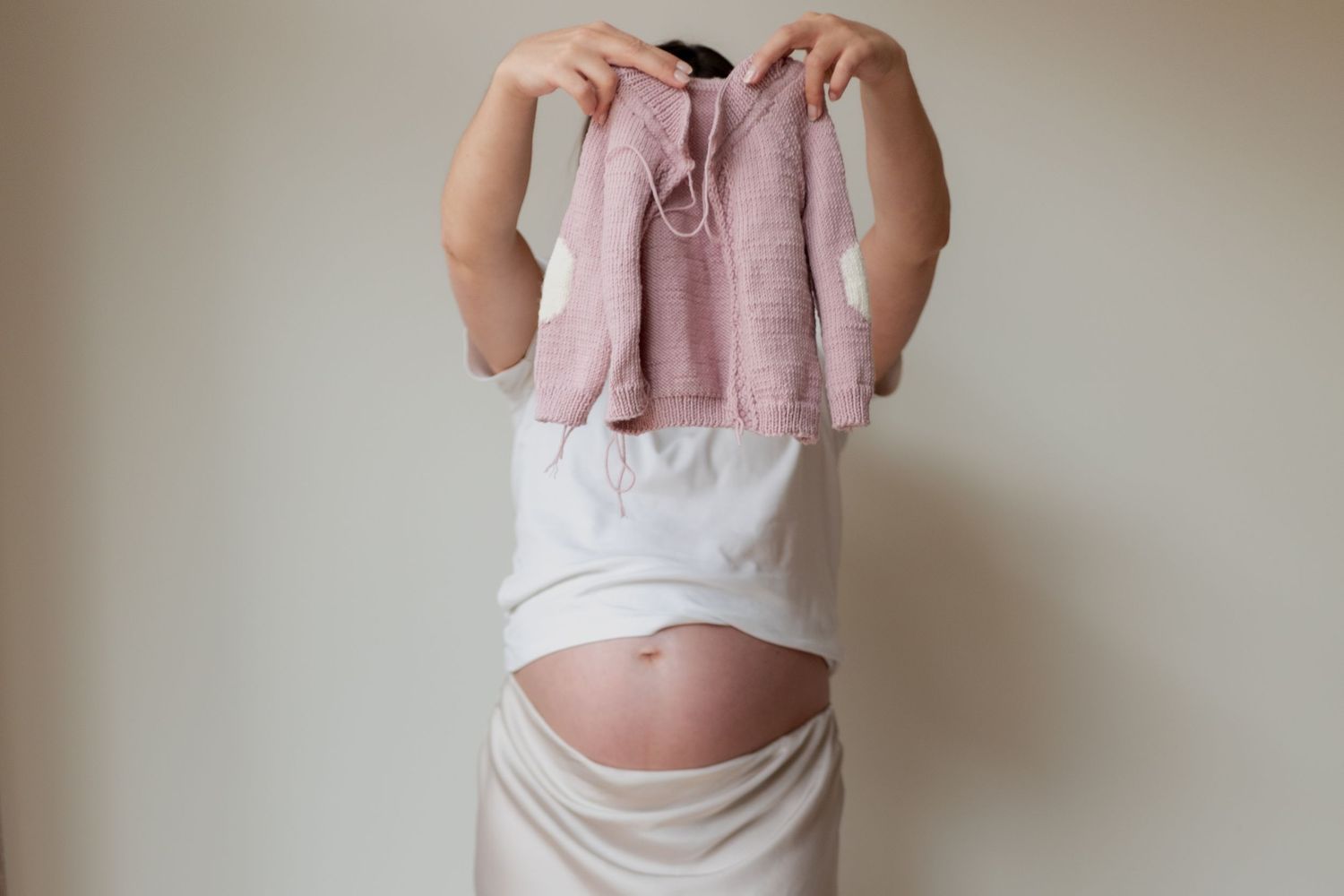 A pregnant belly is shown with hands holding up a dusty pink baby cardigan against a neutral wall.