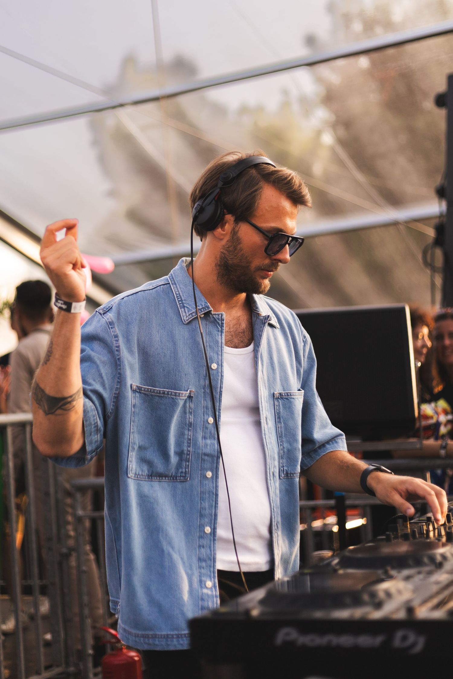DJ wearing denim jacket and sunglasses performing at an outdoor event during golden hour.