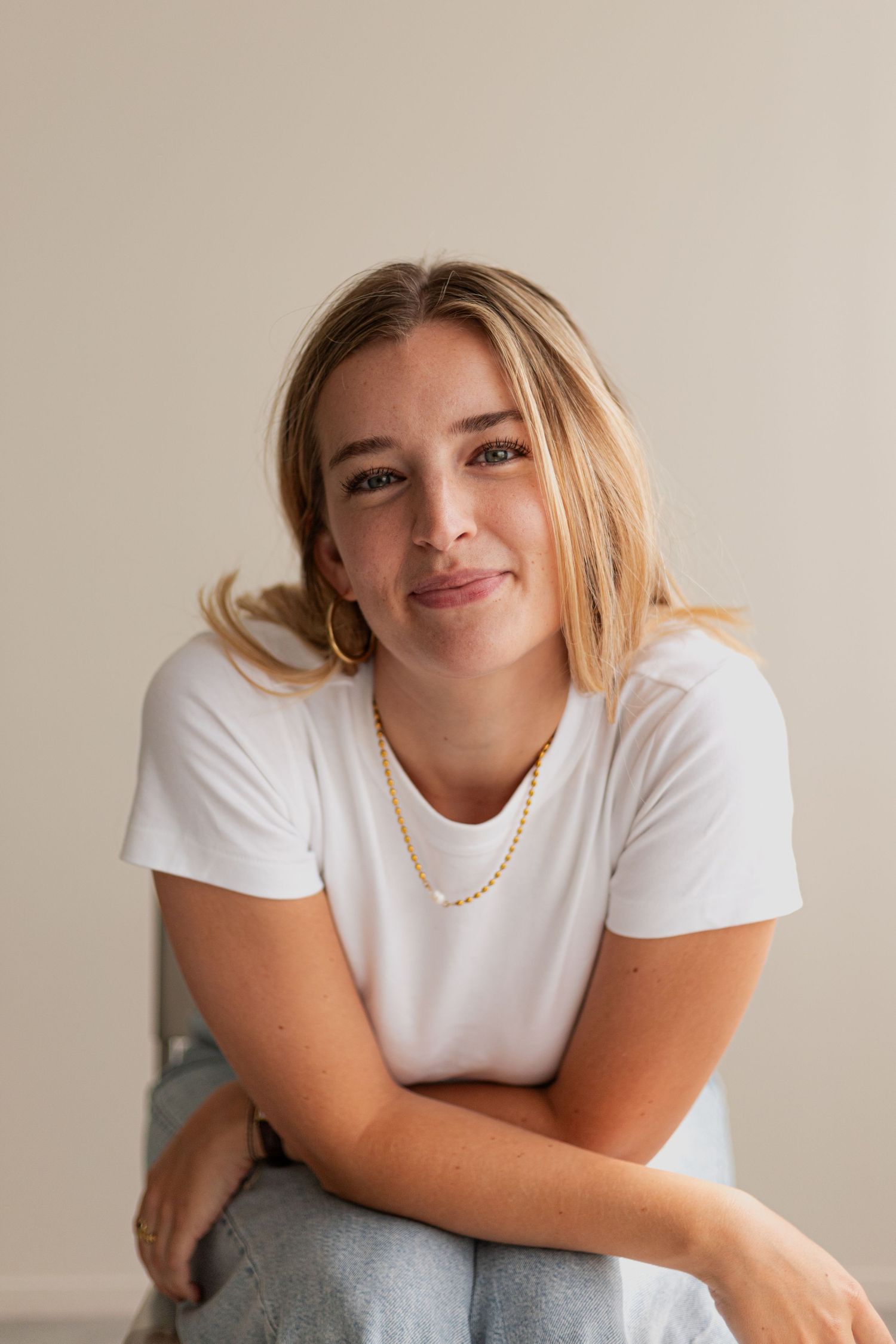 Model in white t-shirt and pearl necklace poses against beige wall in casual portrait series.