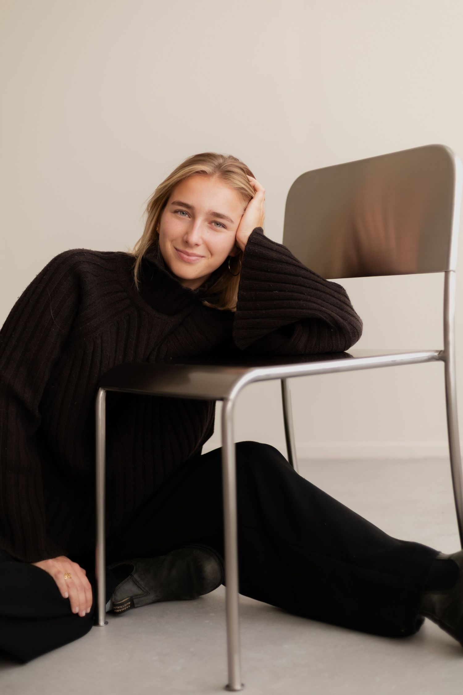 Person in black clothing lying on floor next to modern chair in minimalist setting.