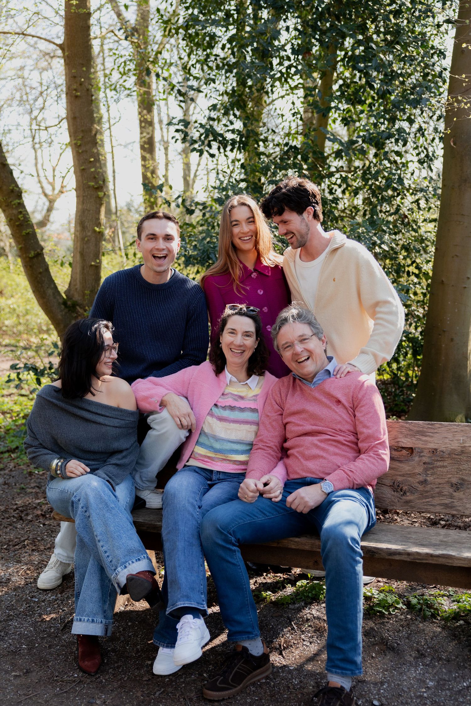 A joyful group of five friends laughing together on a wooden bench in a sunny forest setting.