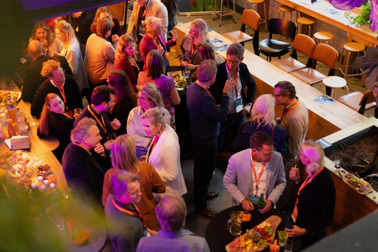 Guests mingle and socialize at a lively indoor event with purple and orange ambient lighting.