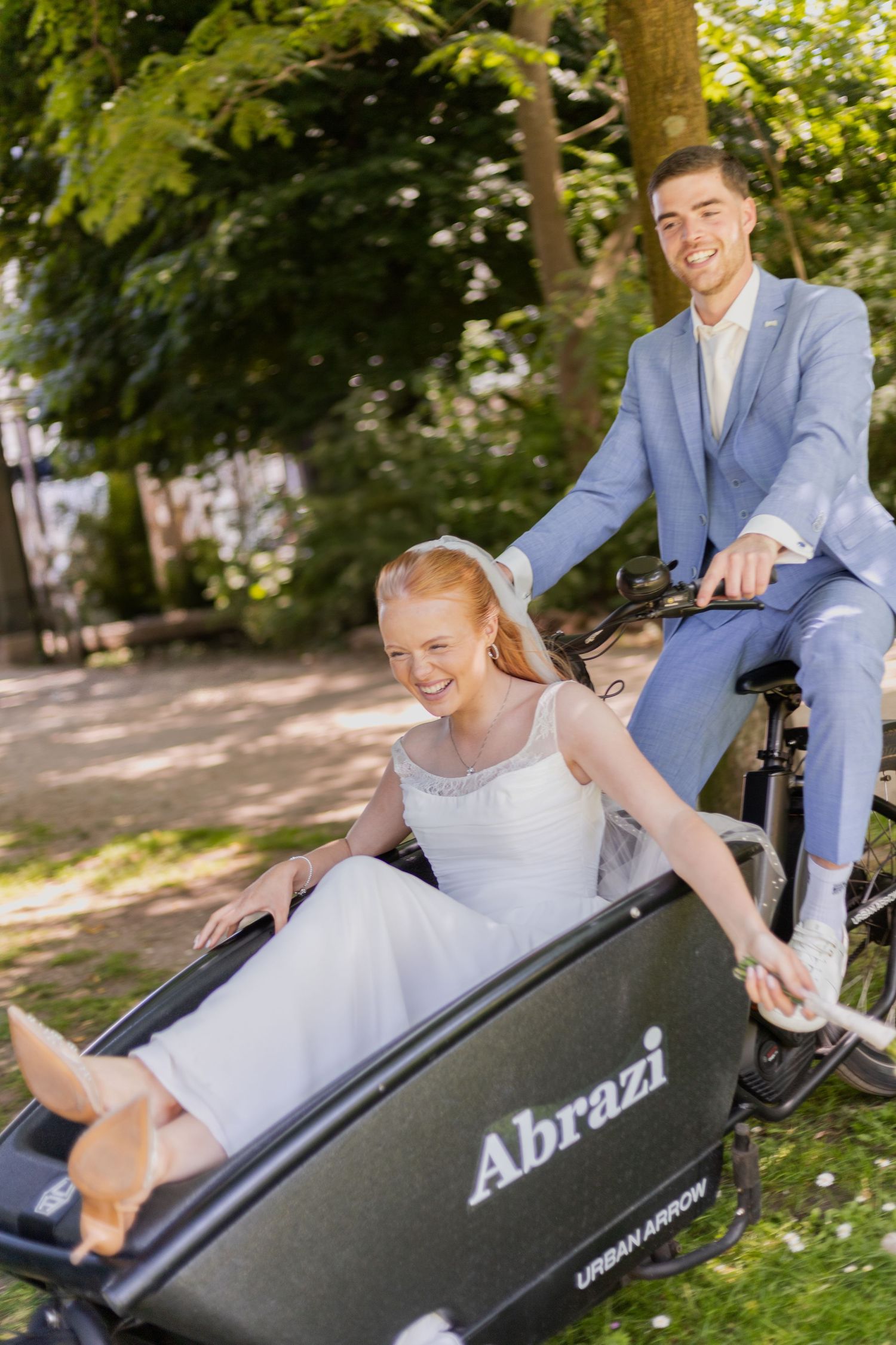 A couple shares a joyful moment in a cargo bike labeled Abrazi during an outdoor photo session in a park setting.