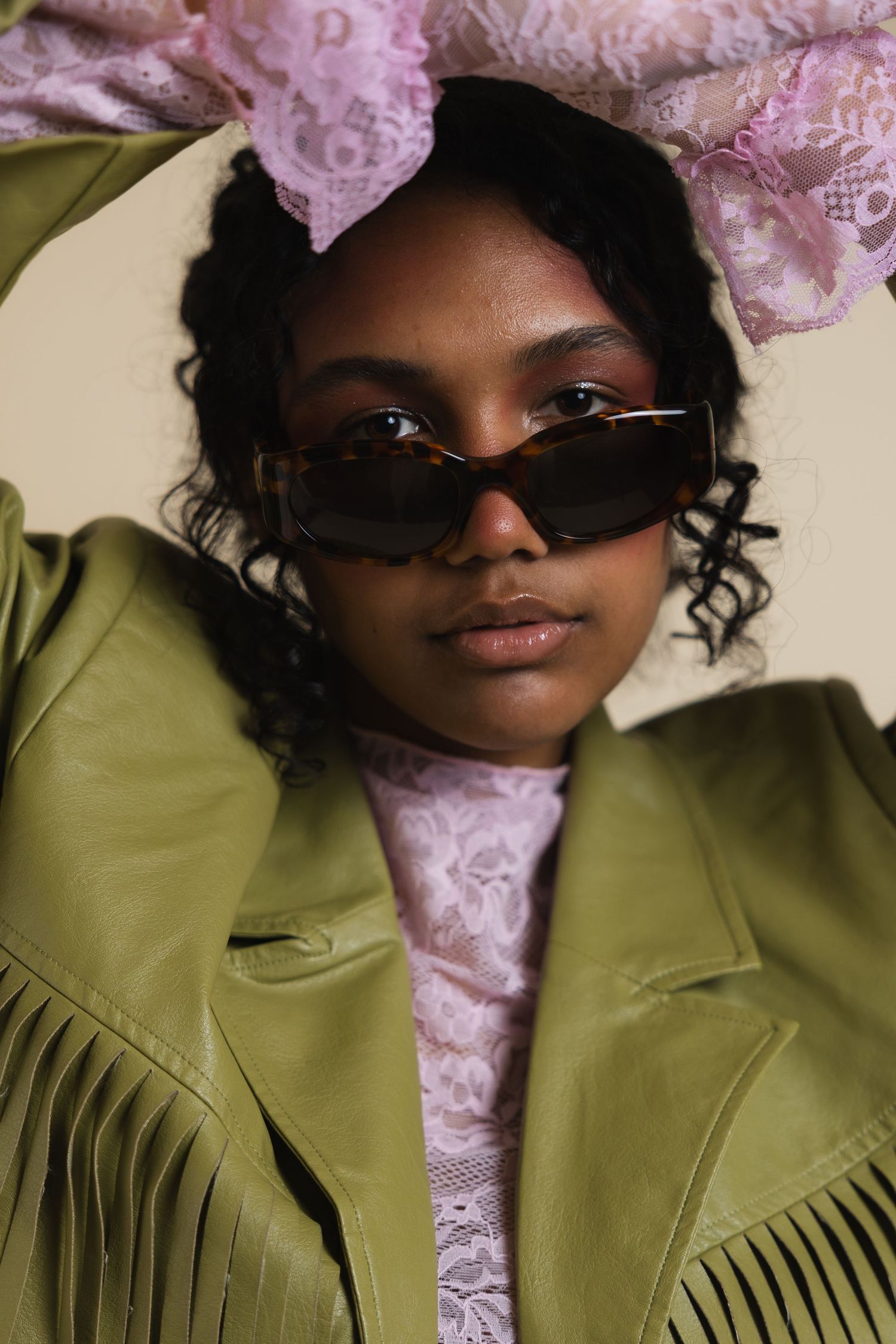 Young woman in green fringe jacket and pink lace outfit wearing retro sunglasses and floral headpiece.