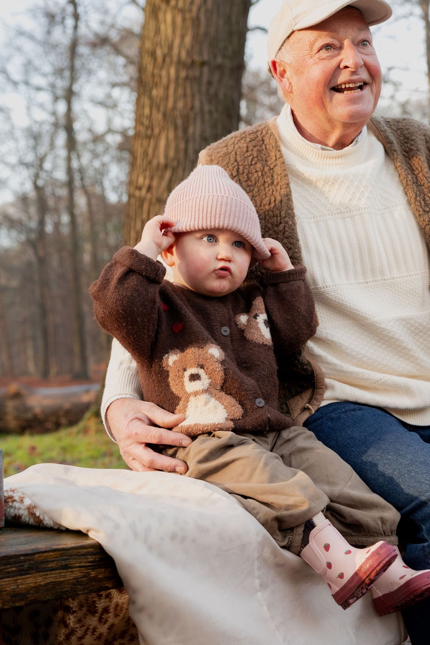 A person wearing a white sweater and brown coat sits on a log outdoors while holding a small child in winter clothing.