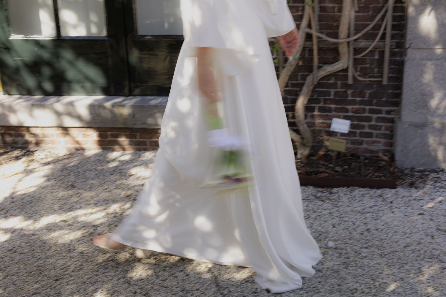 Bride in flowing white dress walking through dappled sunlight in garden setting.