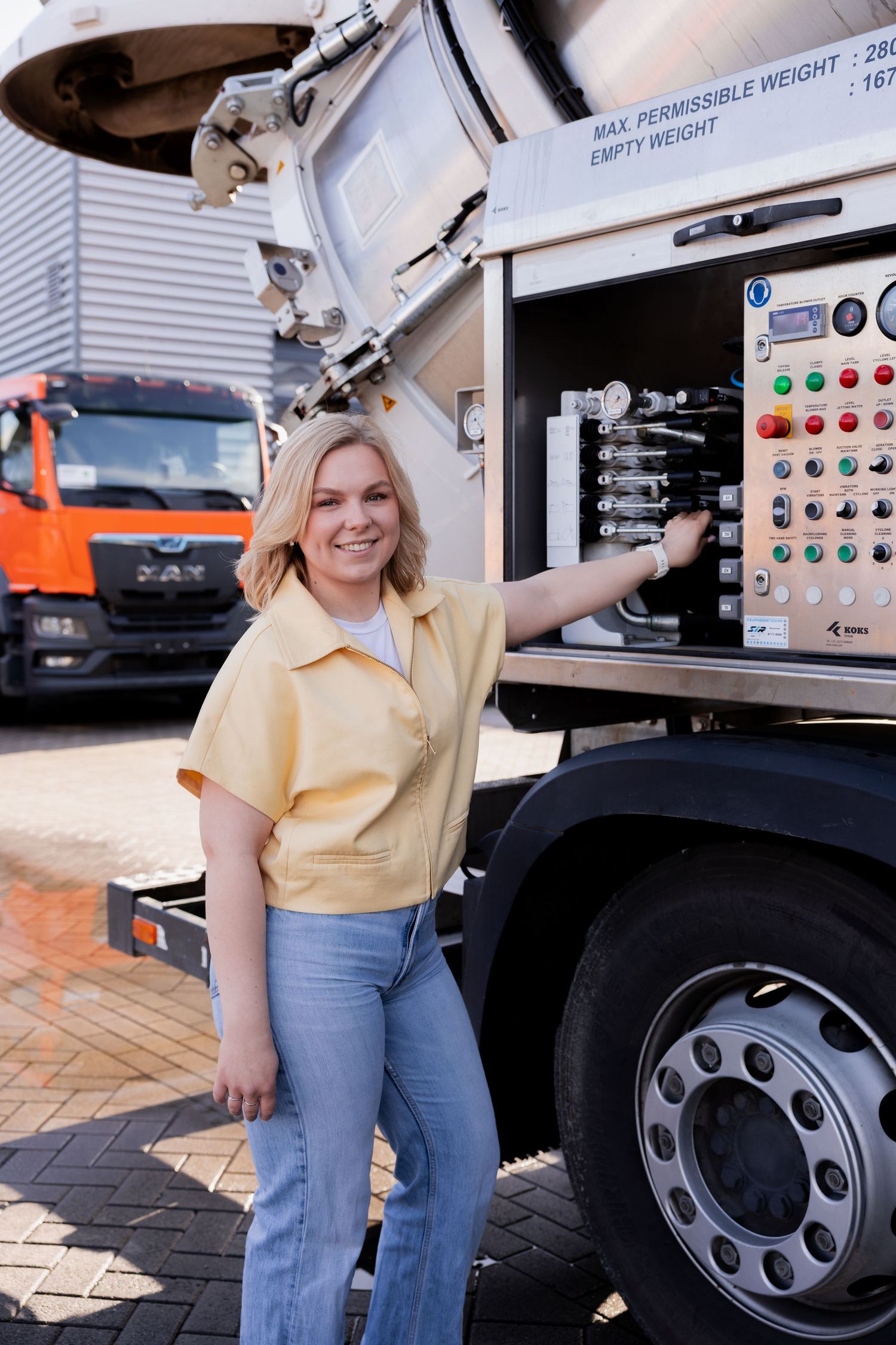 Smiling blonde woman in yellow top and jeans standing beside industrial truck control panel outdoors.