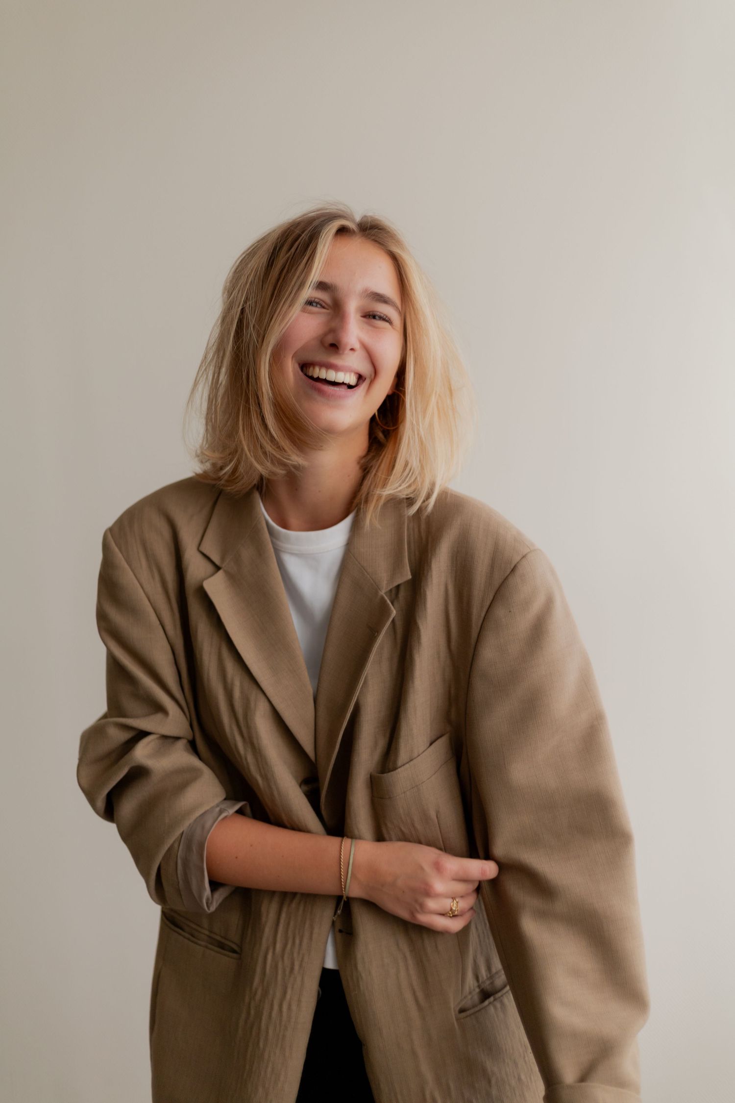 Person in oversized beige blazer laughing and posing against neutral background in casual photo series.