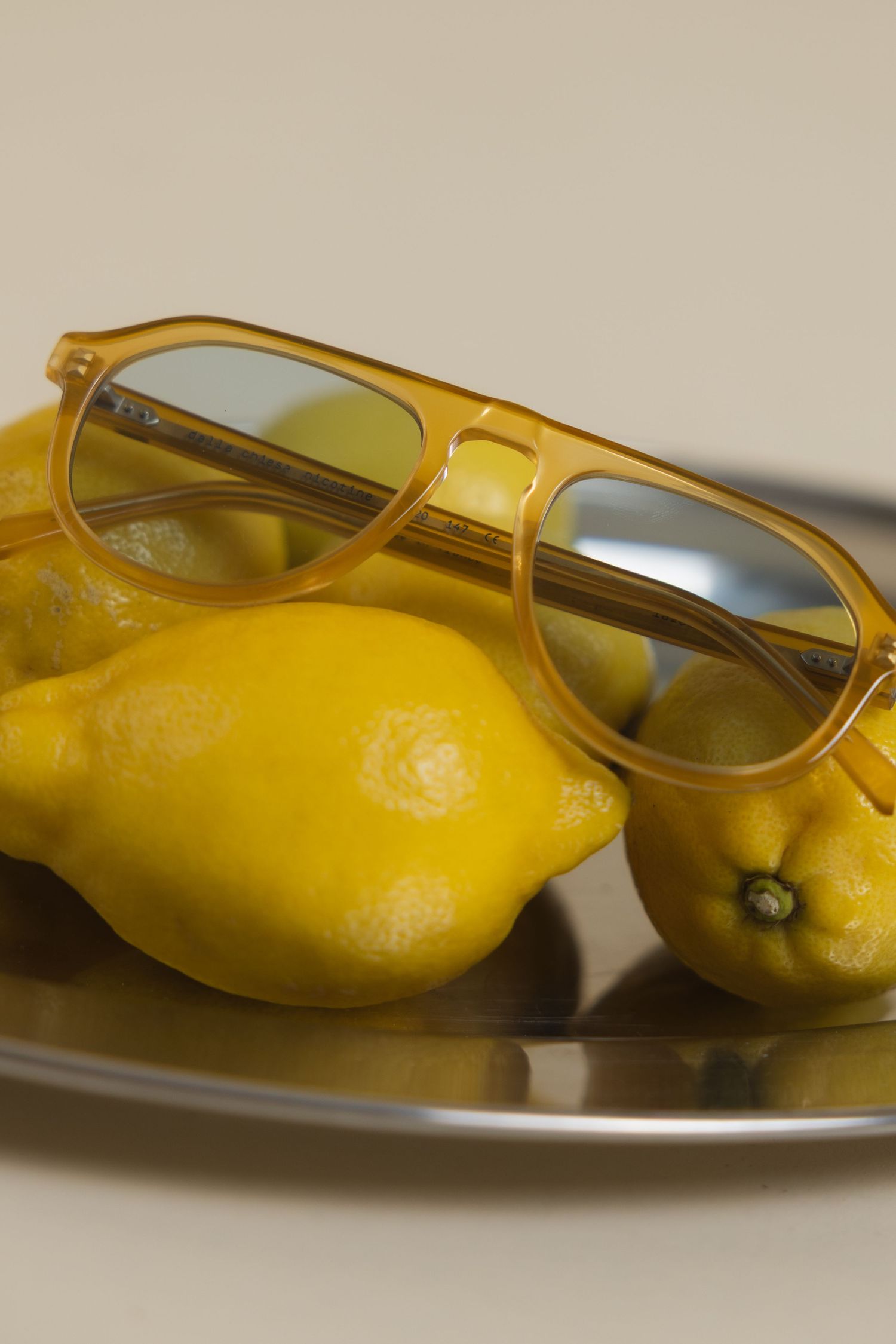 Yellow aviator sunglasses resting on fresh lemons arranged on a silver tray against a neutral background.