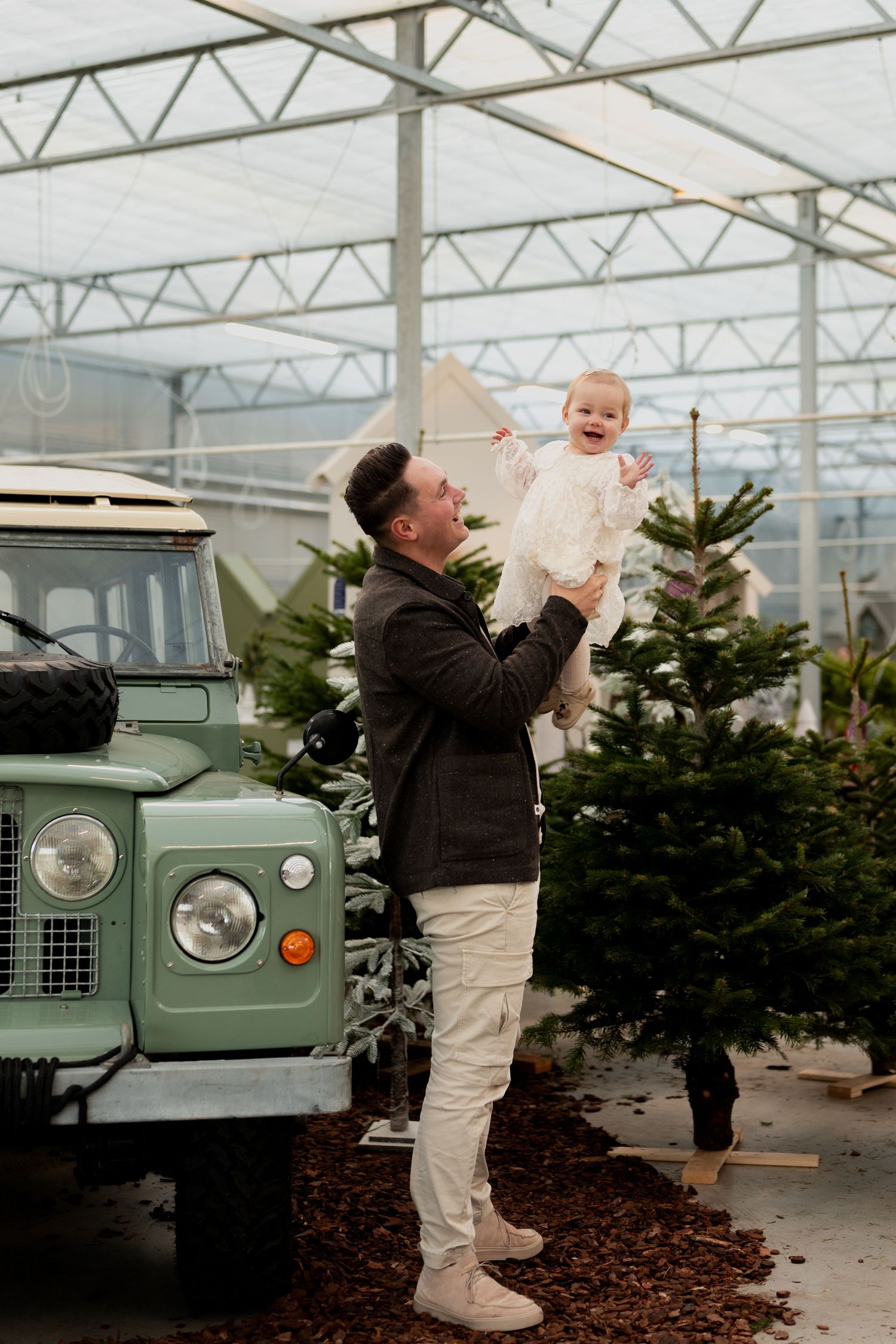 A series of images showing someone lifting a baby near a vintage green Land Rover and Christmas trees in a greenhouse.