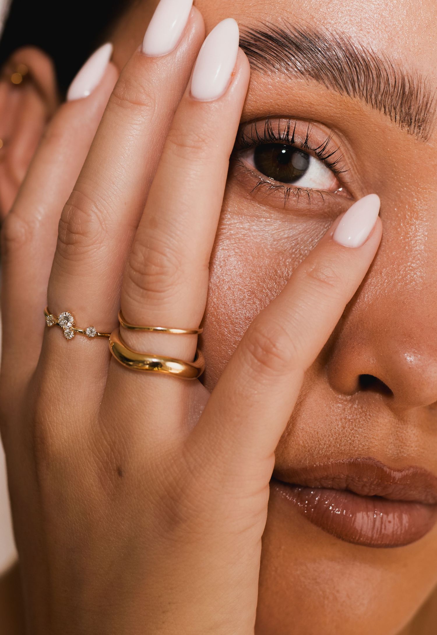 Close-up beauty shot showing white manicured nails, delicate gold rings, and soft neutral makeup details.
