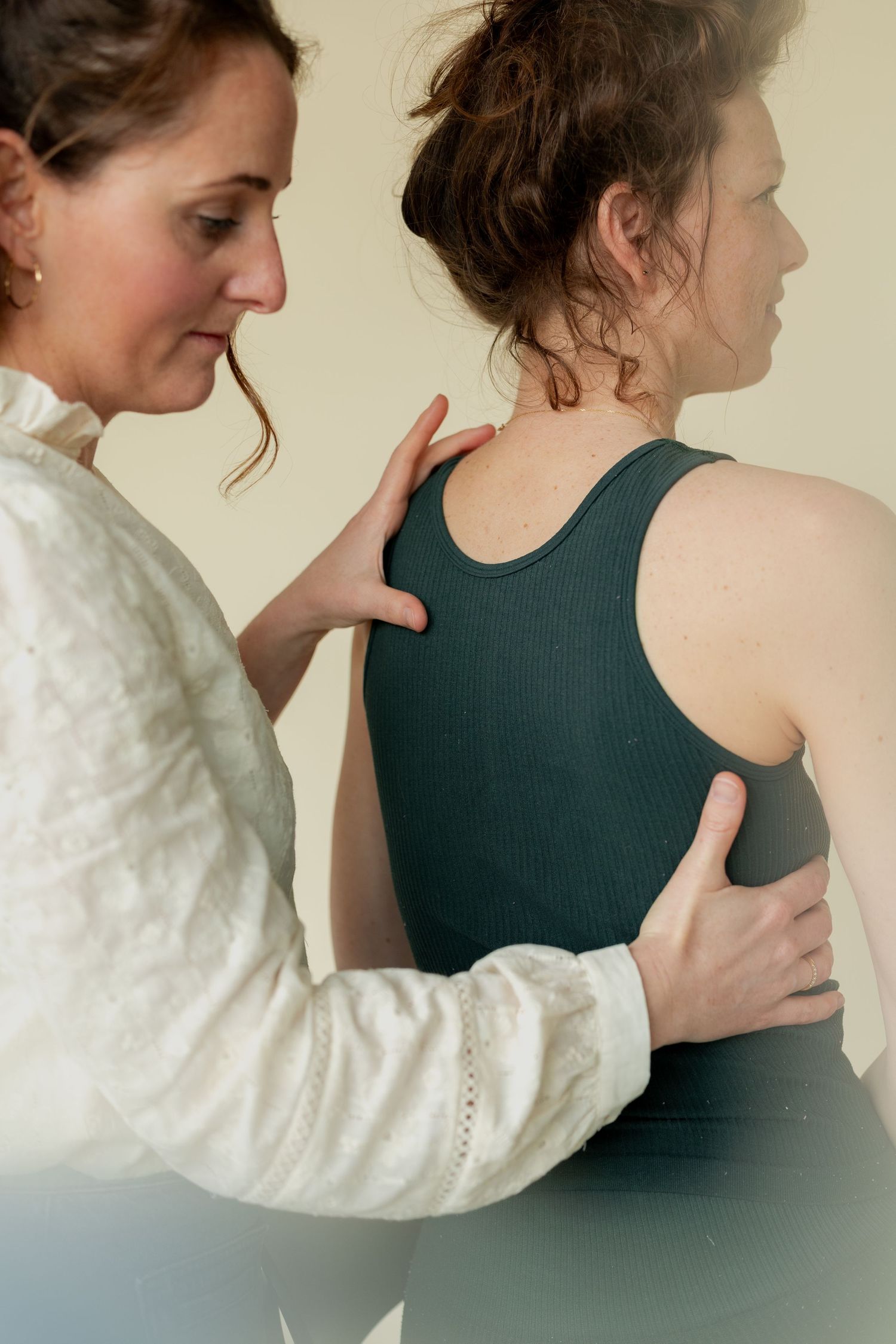 A therapist in a white blouse assesses a woman's posture by placing hands on her back and shoulder.