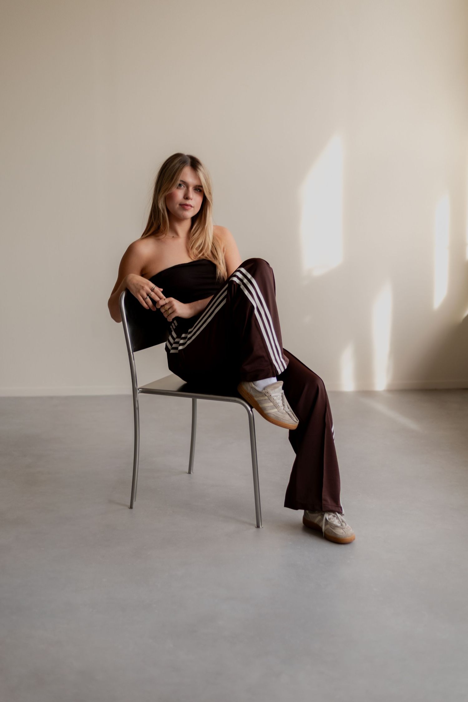 A person in dark clothing poses artistically on a minimalist chair against a light-colored wall in a studio setting.