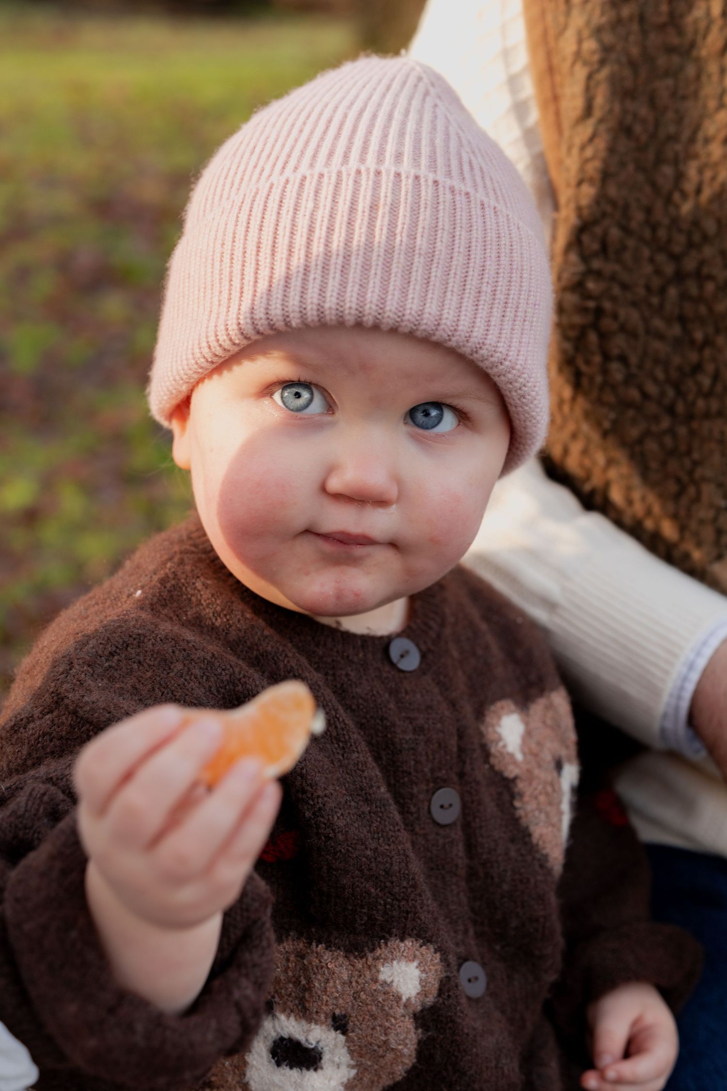 Close up view of a baby wearing a pink knit hat and brown sweater during an autumn day outdoors.