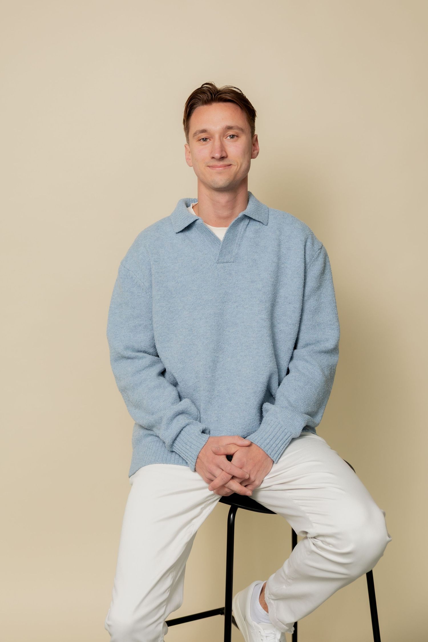 Person in light blue sweatshirt and white pants sits casually on black stool against beige background.