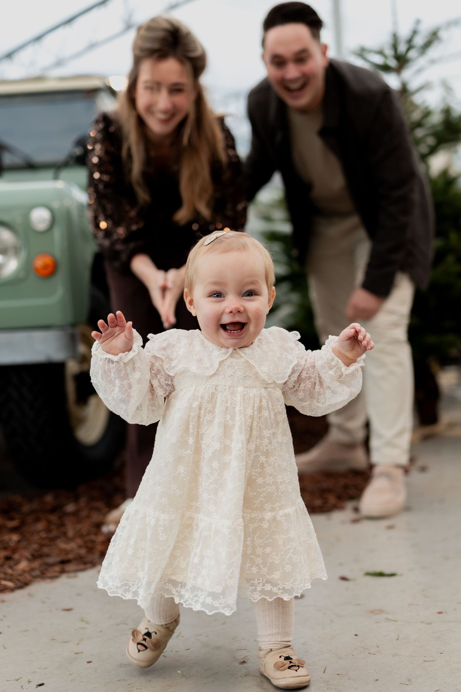 A baby in a white lace dress laughs joyfully while taking first steps on a concrete path.