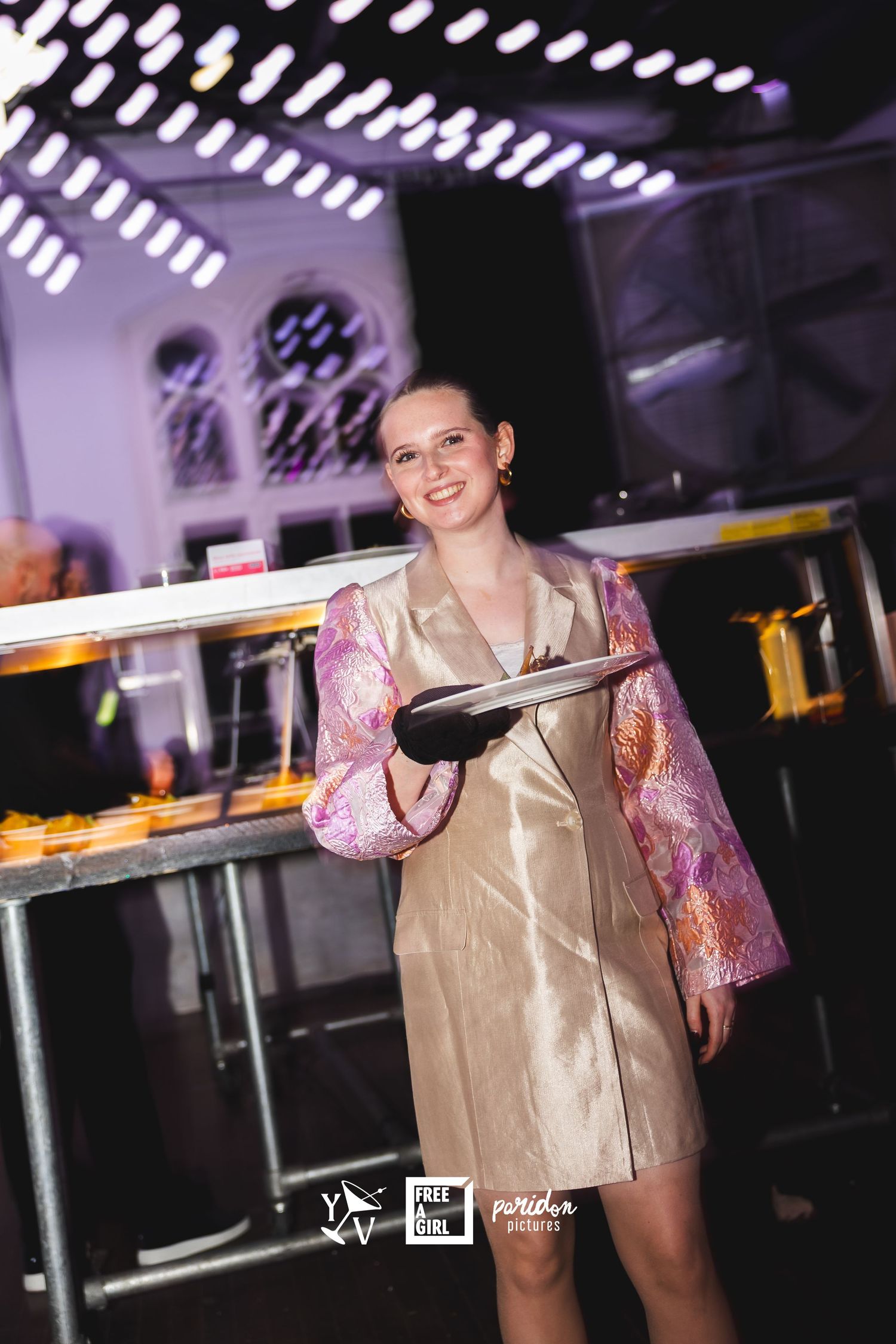 Server holding a tray in a venue wearing a beige dress with pink patterned sleeves in a dimly lit space.