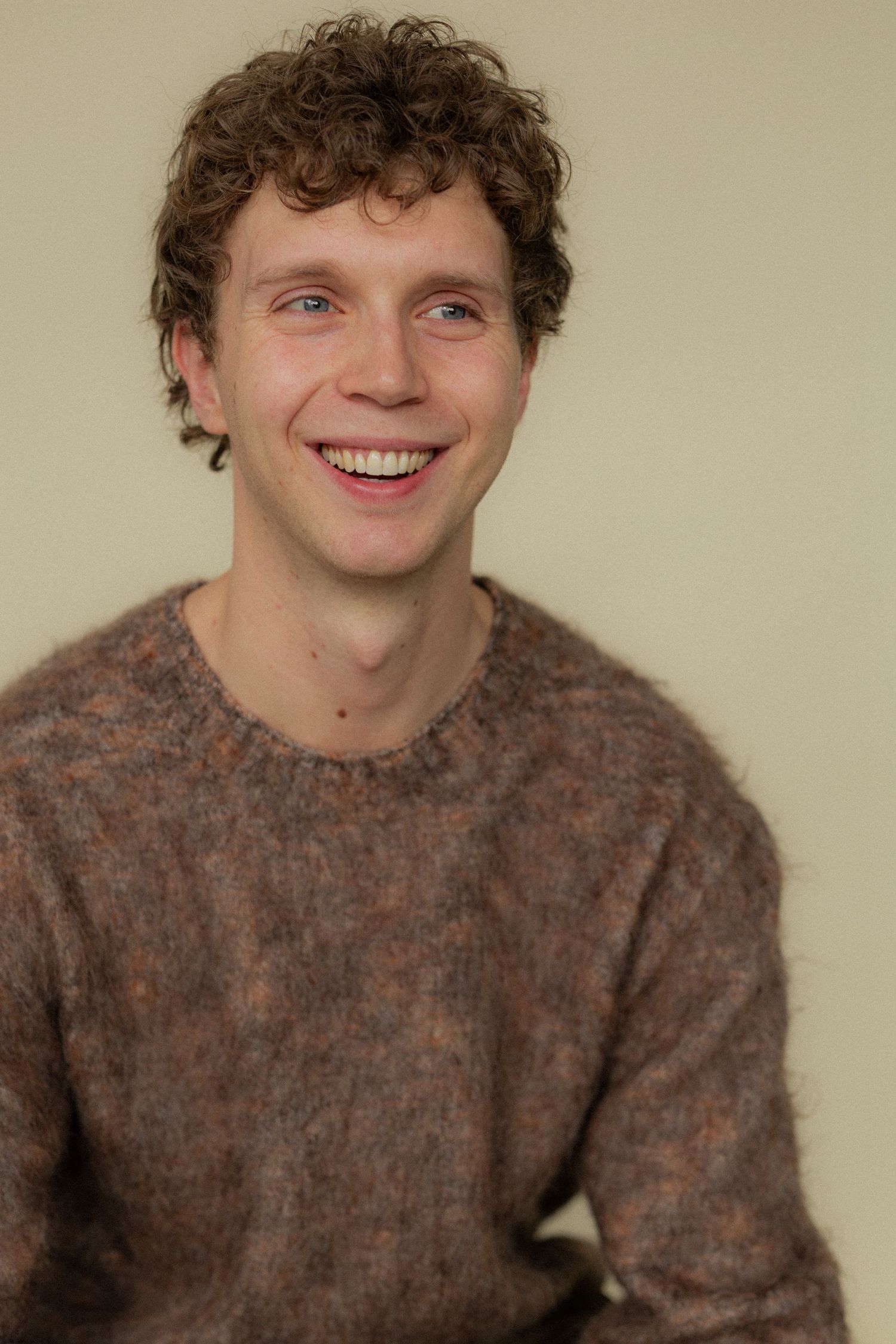 Portrait of someone in a brown textured sweater with curly hair and a warm smile against a beige background.