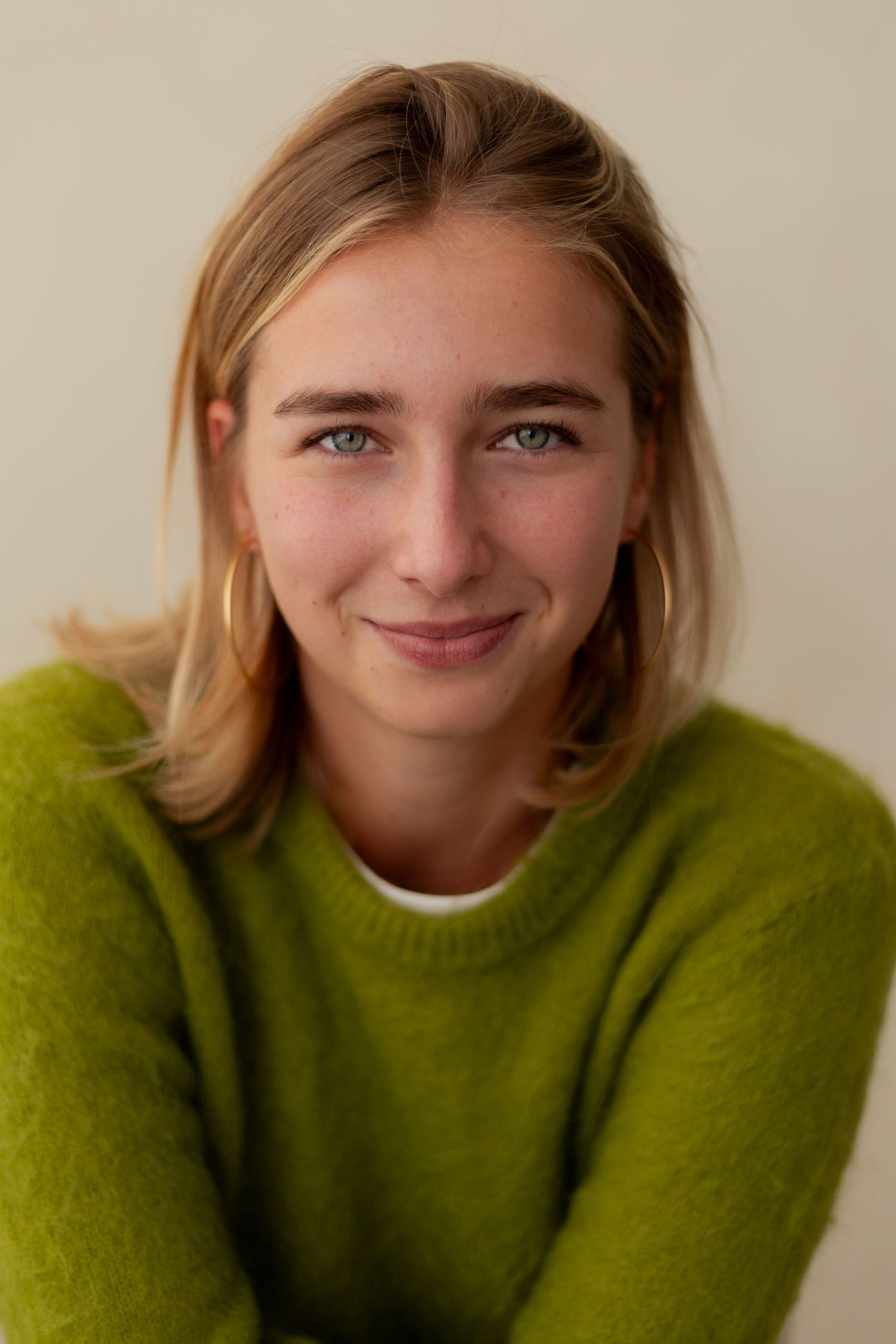 Young blonde woman with blue eyes smiling warmly, wearing a green fuzzy sweater and gold hoop earrings.