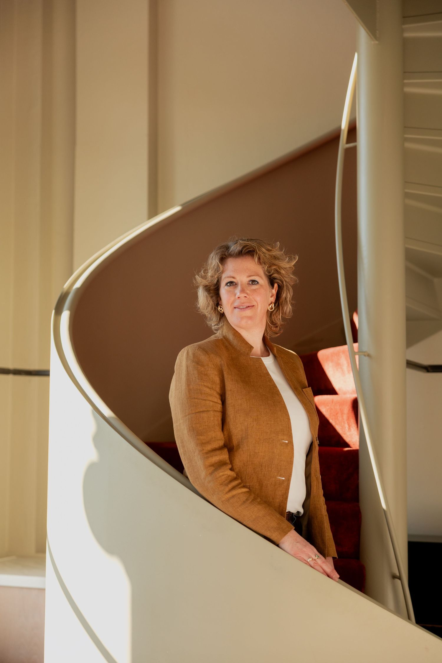 Woman in brown blazer poses elegantly on a cream spiral staircase in a sunlit interior space.