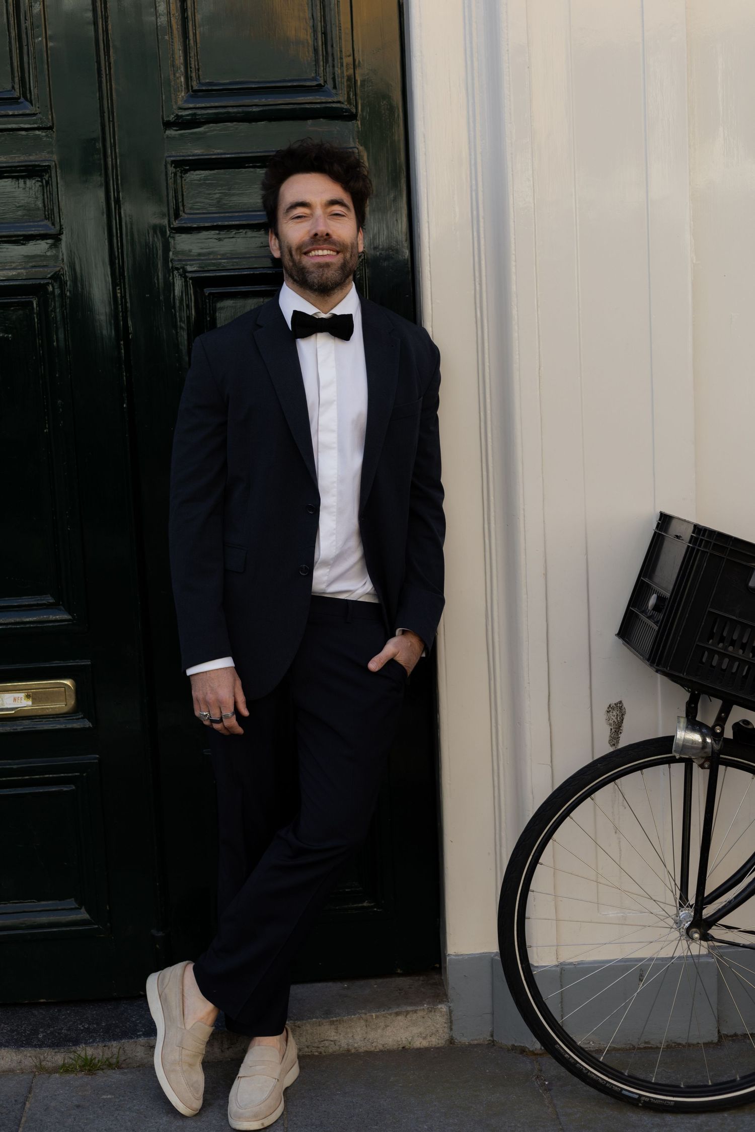 Person in formal black tuxedo poses stylishly against a white wall next to a bicycle.