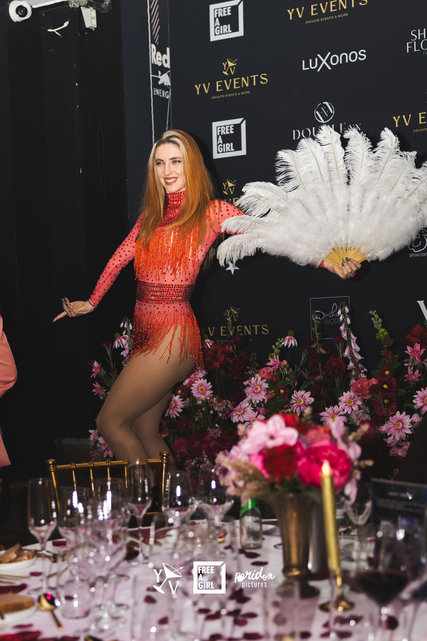 Elegant event setup with floral centerpieces, wine glasses, and stage performance featuring red costume and white feathers.