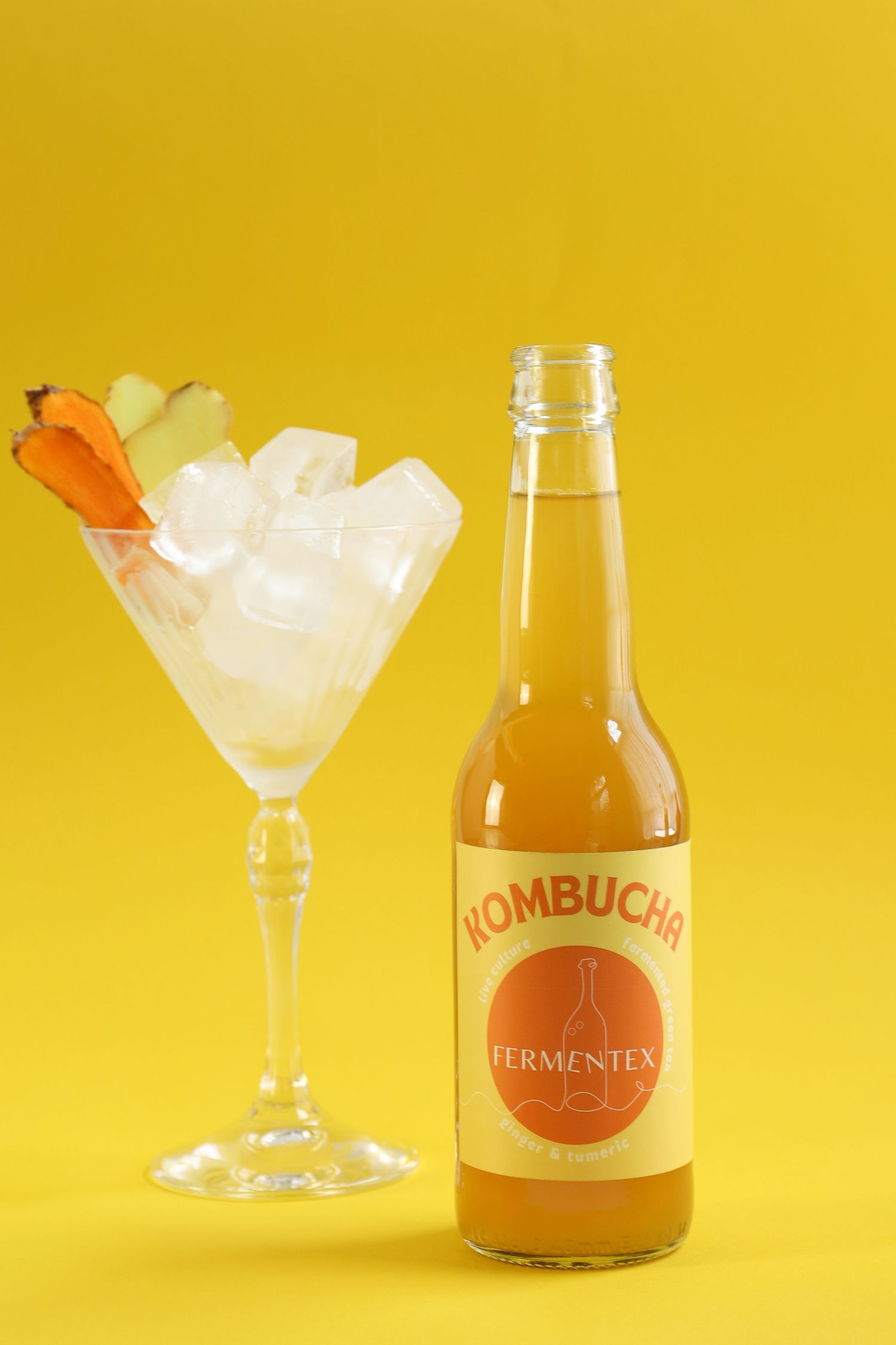 Kombucha bottle and cocktail glass with ice against vibrant yellow background.