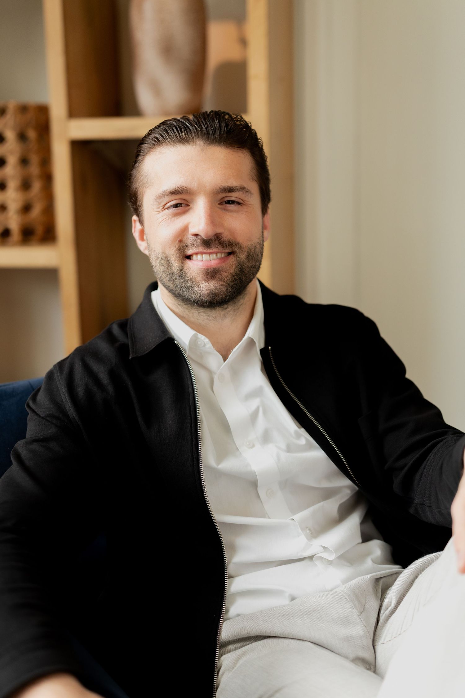 Smiling bearded man in black jacket and white shirt sitting relaxed on a sofa in a modern interior.