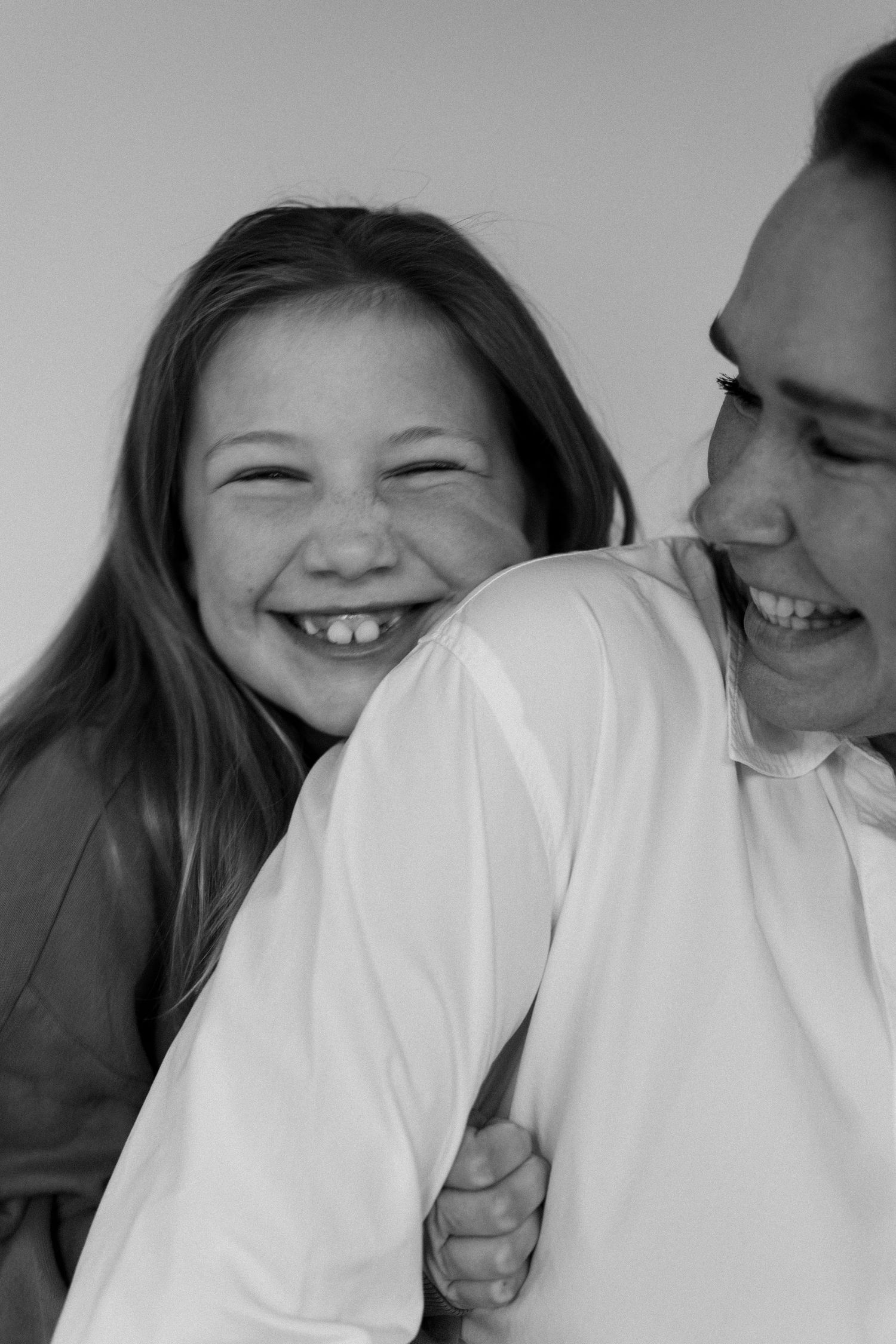 Black and white photo of a laughing girl hugging an adult from behind, both smiling joyfully.