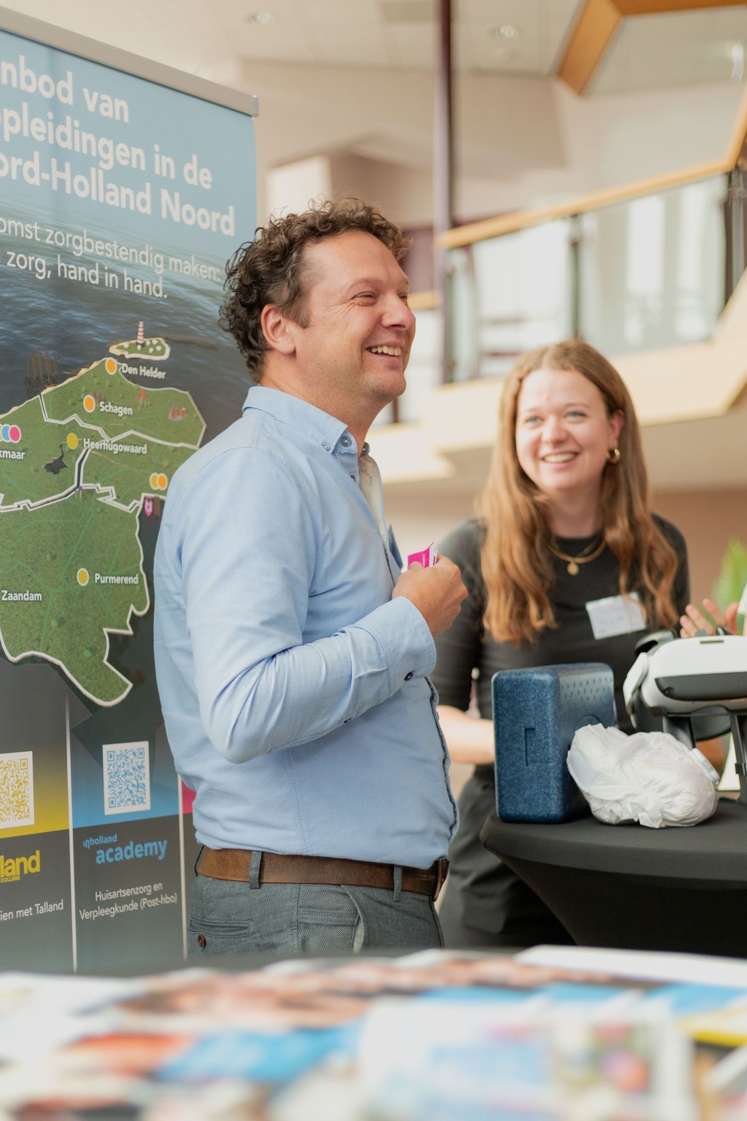 Two people having a cheerful interaction at an information desk with a map of Noord-Holland displayed on the wall.