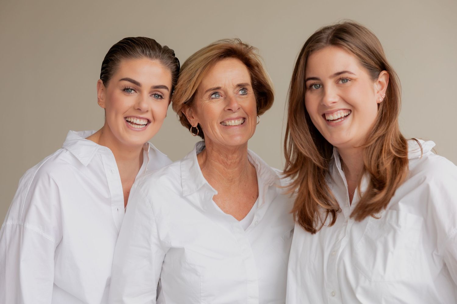 Three women in matching white button-down shirts pose together for a portrait against a neutral background.