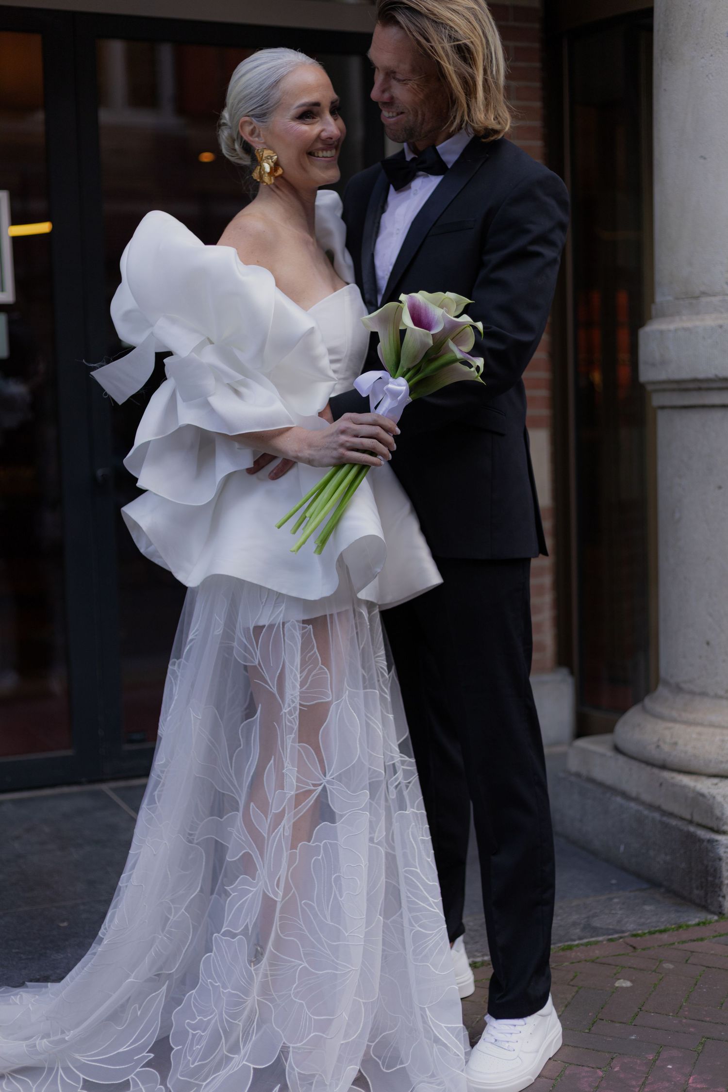 Modern bridal look featuring a structured white dress with calla lilies outside a building.