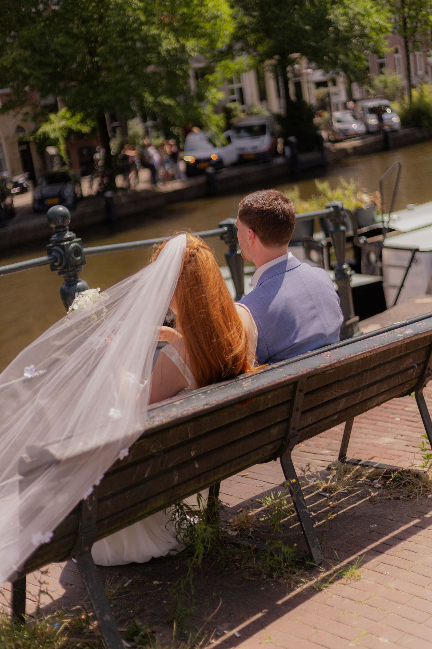 A wedding couple sits on a wooden bench along a tree-lined canal on their special day.
