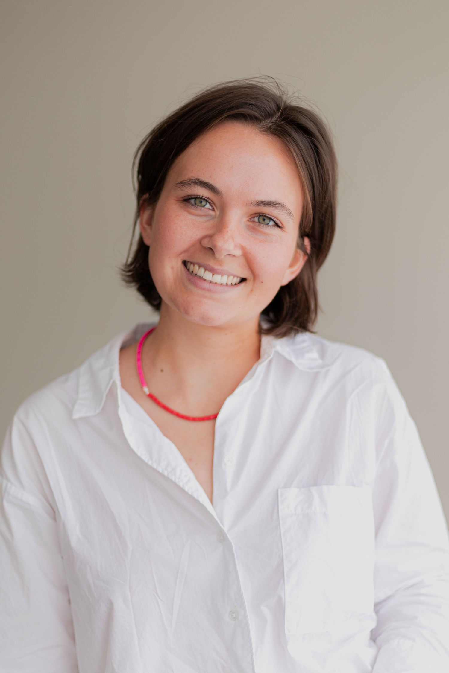 Lachende jonge vrouw met kort bruin haar in wit shirt en roze kralenketting, tegen een grijze achtergrond.
